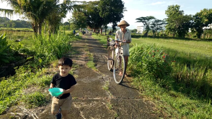 A young boy in a black shirt and tan shorts holds a turquoise bowl while walking down a rural path, an elderly man pushes a bicycle beside tall grass and trees in a sunny field.