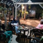 Three adults seated in a circular arrangement around a small table with drinks and snacks, chatting under a wooden pergola in a nighttime garden setting.