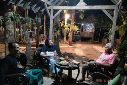 Three adults seated in a circular arrangement around a small table with drinks and snacks, chatting under a wooden pergola in a nighttime garden setting.