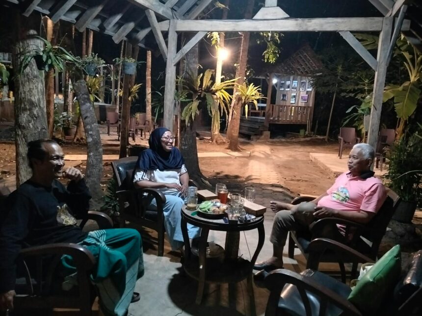 Three adults seated in a circular arrangement around a small table with drinks and snacks, chatting under a wooden pergola in a nighttime garden setting.