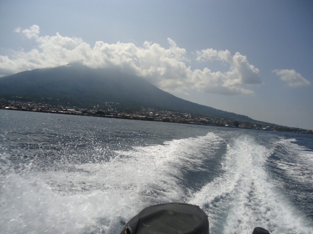 WhatsApp Image 2026-09-08 at 143653 - Mabur.co Boating scene: white wake and spray in the foreground, with a coastal town along the shore and a green mountain under a partly cloudy sky.
