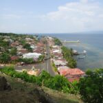 Coastal town built on a hillside with a main road running toward the sea, red-roofed houses and palm trees lining the street.