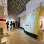 Large textile artwork hangs from the ceiling in a gallery, featuring silhouettes of people in motion with yellow and dark blue borders as visitors look on.
