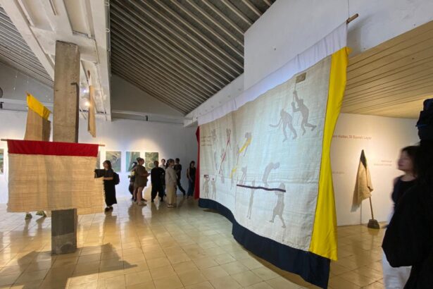 Large textile artwork hangs from the ceiling in a gallery, featuring silhouettes of people in motion with yellow and dark blue borders as visitors look on.