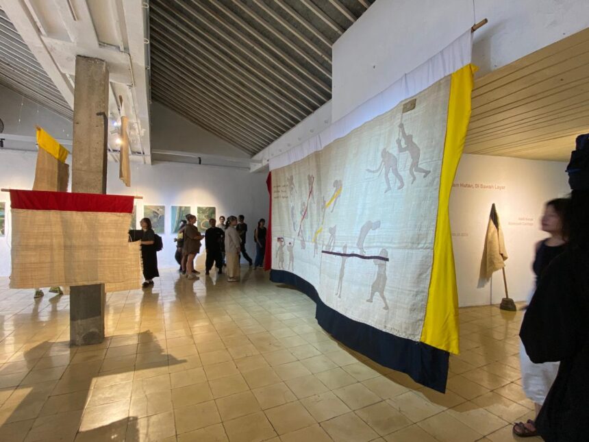 Large textile artwork hangs from the ceiling in a gallery, featuring silhouettes of people in motion with yellow and dark blue borders as visitors look on.