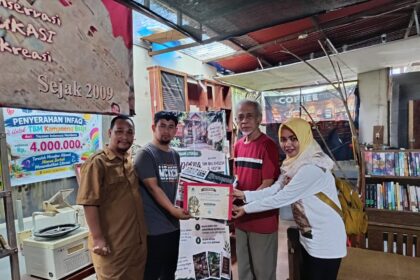 Group of four people posing as one person receives a certificate in a small library or bookshop setting, with banners and books in the background.