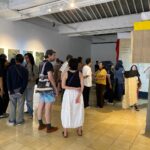 Group of people in a modern gallery looking at wall-mounted photos; a beige fabric sculpture drapes from a concrete pillar nearby.