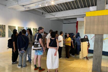Group of people in a modern gallery looking at wall-mounted photos; a beige fabric sculpture drapes from a concrete pillar nearby.