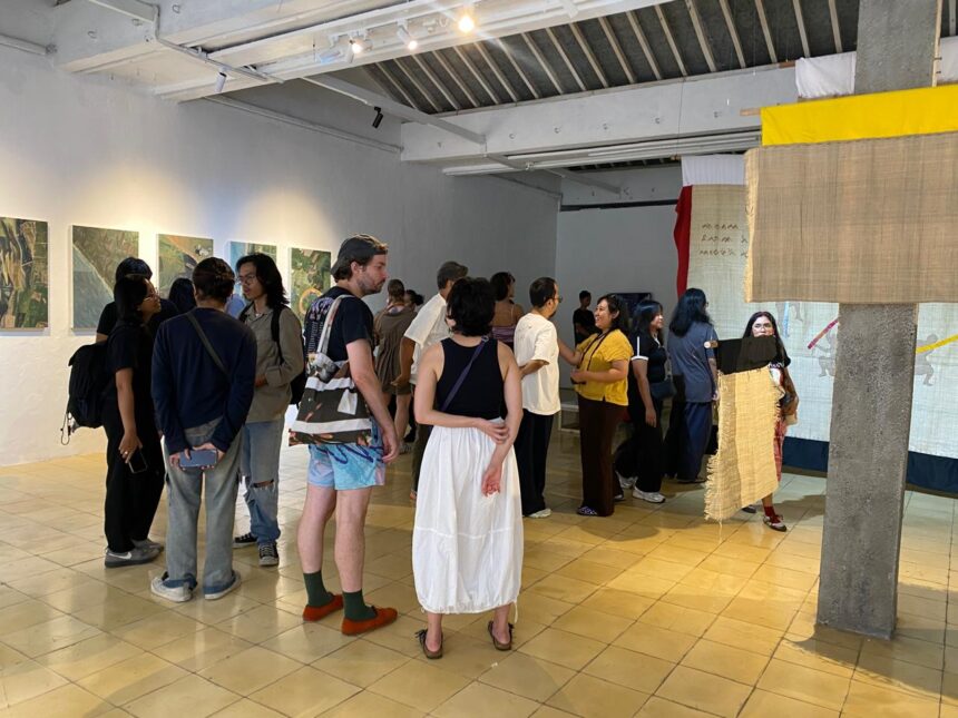 Group of people in a modern gallery looking at wall-mounted photos; a beige fabric sculpture drapes from a concrete pillar nearby.