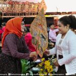 Two women exchange a painted decorative fan during a cultural ceremony, with musicians and flower decorations in the background.