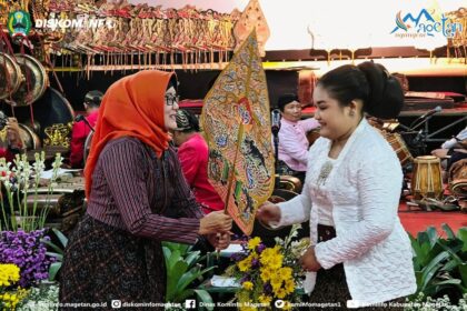Two women exchange a painted decorative fan during a cultural ceremony, with musicians and flower decorations in the background.