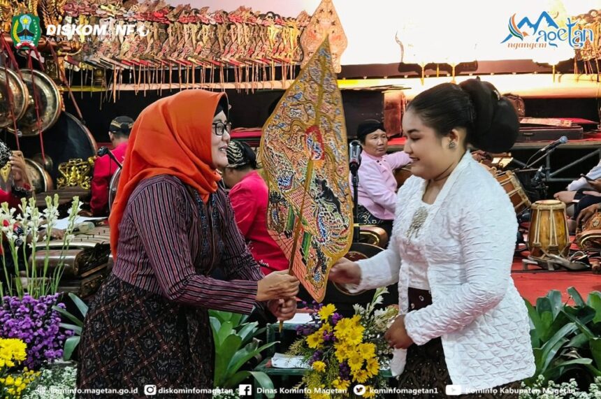Two women exchange a painted decorative fan during a cultural ceremony, with musicians and flower decorations in the background.