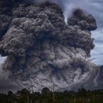 Massive ash plume erupts from a volcano, dark clouds billowing into a gray sky behind a rural landscape.