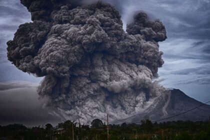 Massive ash plume erupts from a volcano, dark clouds billowing into a gray sky behind a rural landscape.