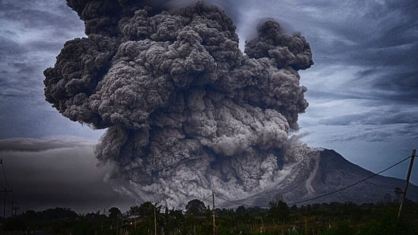 Massive ash plume erupts from a volcano, dark clouds billowing into a gray sky behind a rural landscape.