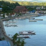 Coastal harbor with several small boats anchored by a stone quay and a row of colorful buildings along the waterfront-palm trees in the foreground and hills in the background.