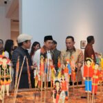 Group of people in traditional and modern clothing viewing a tabletop display of colorful Indonesian puppets on sticks at an exhibition hall.
