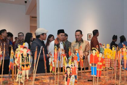 Group of people in traditional and modern clothing viewing a tabletop display of colorful Indonesian puppets on sticks at an exhibition hall.