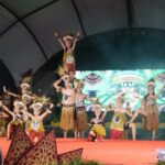 Group of dancers in tribal costumes performing on a red stage; a child is lifted on top of a dancer’s shoulders while others pose with arms raised.