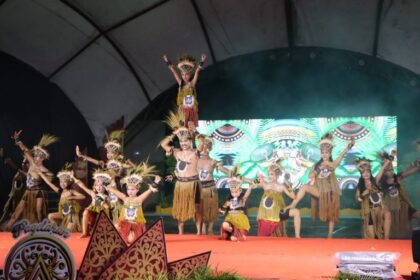 Group of dancers in tribal costumes performing on a red stage; a child is lifted on top of a dancer’s shoulders while others pose with arms raised.