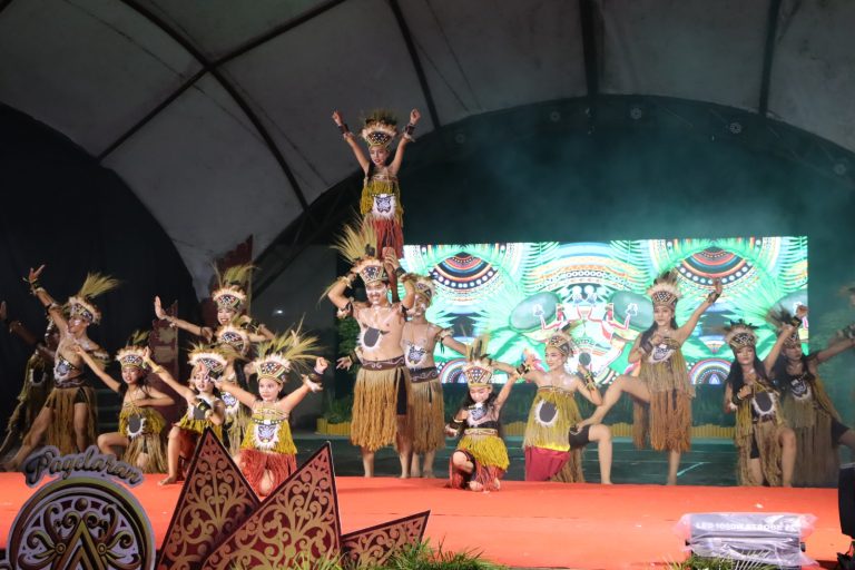 Group of dancers in tribal costumes performing on a red stage; a child is lifted on top of a dancer’s shoulders while others pose with arms raised.