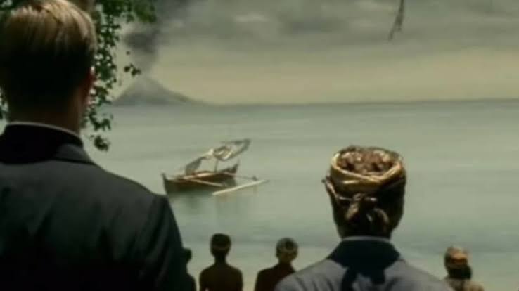 Back view of two men in suits watching a small wooden boat stranded on calm water near shore, with a few onlookers nearby.