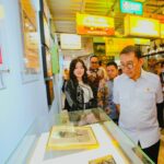 Group of people in batik and white shirts look at a framed photo inside a glass display case at a bright exhibition hall with colorful signs overhead.