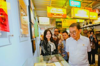 Group of people in batik and white shirts look at a framed photo inside a glass display case at a bright exhibition hall with colorful signs overhead.