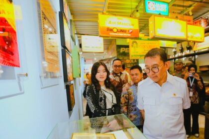 Group of people in batik and white shirts look at a framed photo inside a glass display case at a bright exhibition hall with colorful signs overhead.