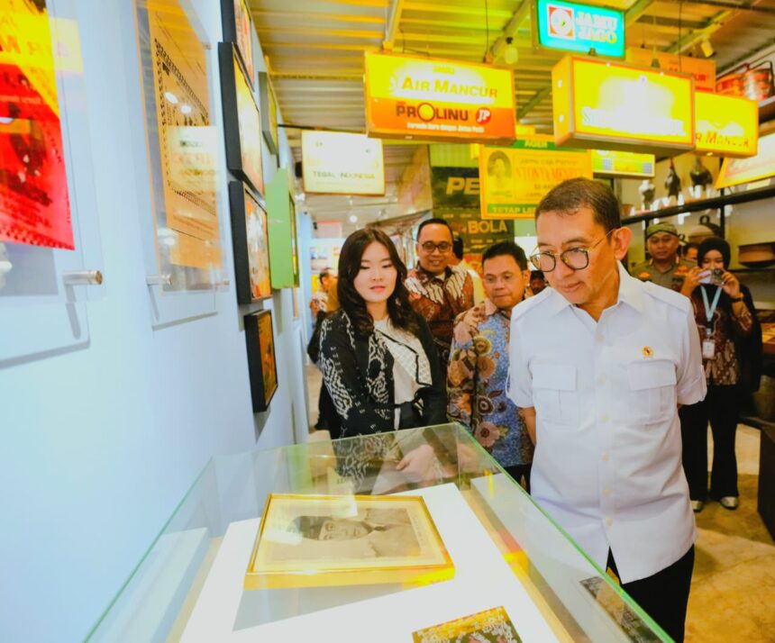 Group of people in batik and white shirts look at a framed photo inside a glass display case at a bright exhibition hall with colorful signs overhead.
