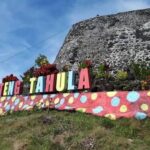 Colorful sign reading 'Benteng Tahula' atop a pink wall with polka dots, in front of a stone fortress and blue sky.