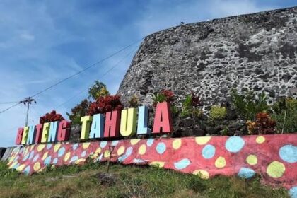 Colorful sign reading 'Benteng Tahula' atop a pink wall with polka dots, in front of a stone fortress and blue sky.