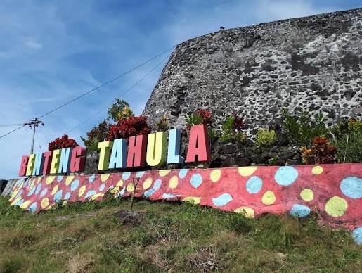 Colorful sign reading 'Benteng Tahula' atop a pink wall with polka dots, in front of a stone fortress and blue sky.