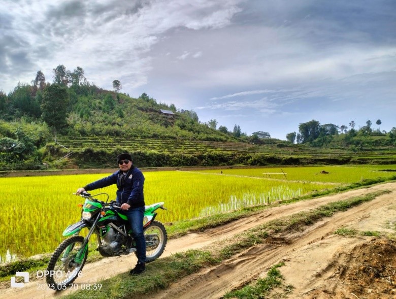 Person on a green dirt bike beside a yellow rice field with terraced hills and a blue sky in the background.
