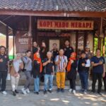 Group of people posing for a photo in front of a rustic coffee stall labeled 'Kopi Madu Merapi', outdoors on a sunny day.