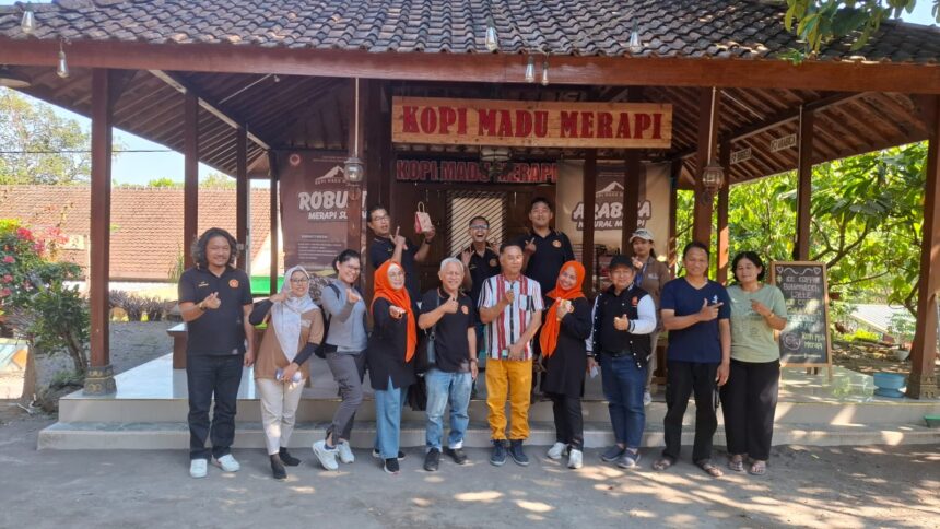 Group of people posing for a photo in front of a rustic coffee stall labeled 'Kopi Madu Merapi', outdoors on a sunny day.