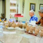 Three men sit around a conference table with numerous carved busts on display, discussing something in a bright room filled with books and art.