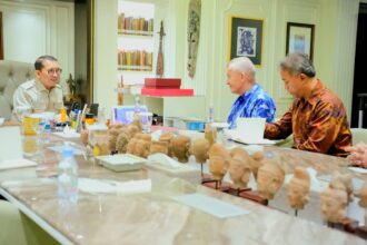 Three men sit around a conference table with numerous carved busts on display, discussing something in a bright room filled with books and art.