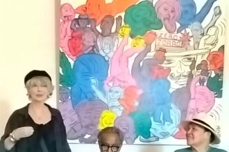 Speaker standing on the left addresses an audience, with two men seated and listening in front of a vivid mural backdrop.