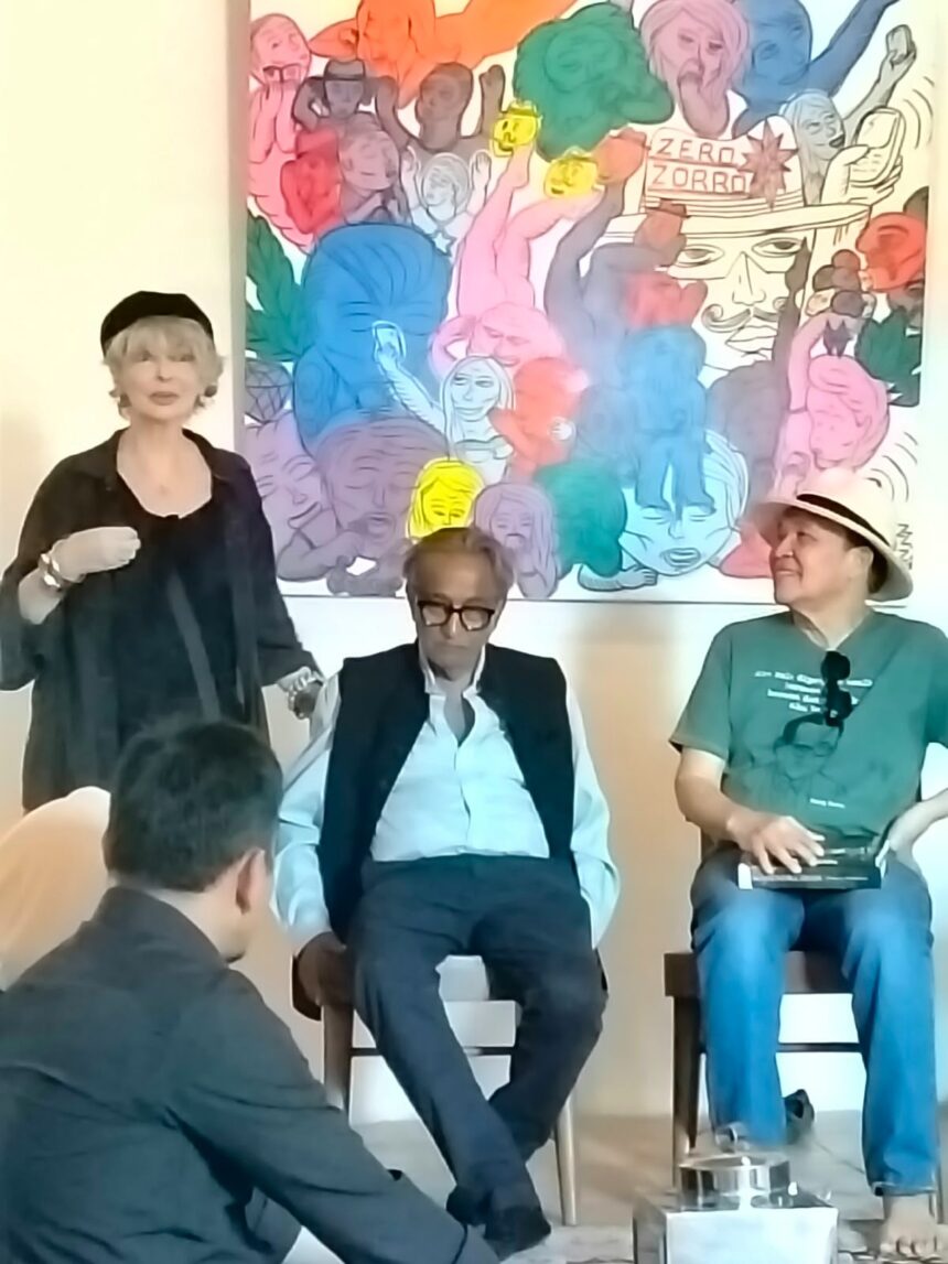 Speaker standing on the left addresses an audience, with two men seated and listening in front of a vivid mural backdrop.