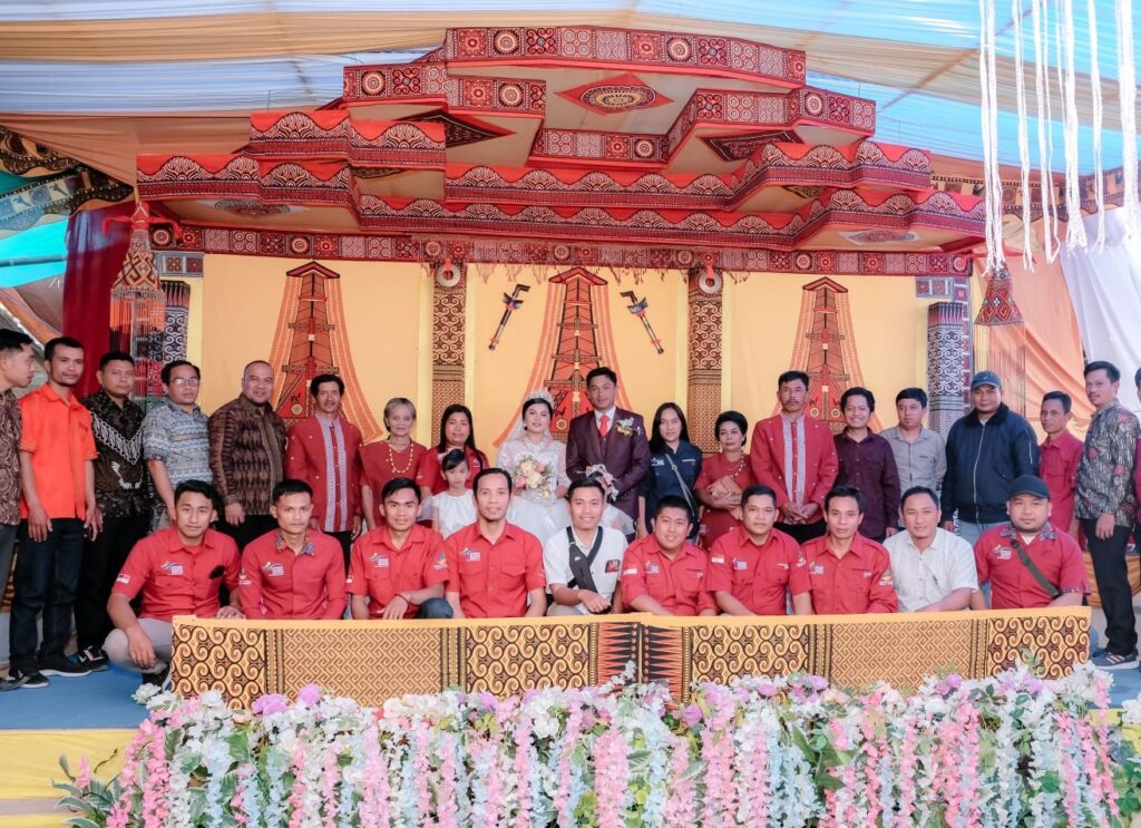 Group photo at a traditional wedding ceremony: bride in a white gown and groom in a suit center, surrounded by family and friends in red attire on a decorated stage with floral garlands and patterned backdrops.