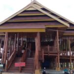 Large wooden stilted building with a grand central staircase, open verandas, and people nearby under a cloudy sky.
