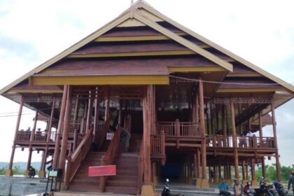 Large wooden stilted building with a grand central staircase, open verandas, and people nearby under a cloudy sky.