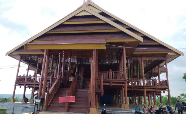 Large wooden stilted building with a grand central staircase, open verandas, and people nearby under a cloudy sky.