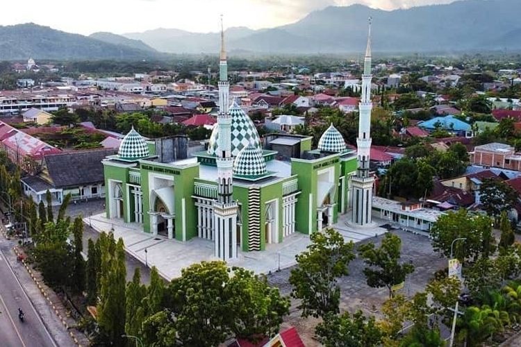Aerial view of a green mosque with white domes and two tall minarets in a town, with mountains in the background.