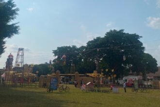Open-air park festival with hay sculptures, hanging decorations, and people strolling under a large tree on a sunny day.