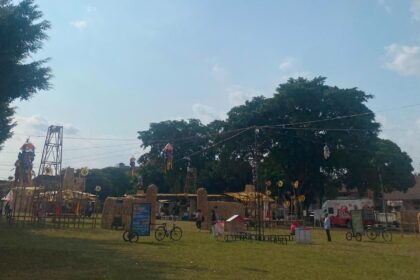 Open-air park festival with hay sculptures, hanging decorations, and people strolling under a large tree on a sunny day.