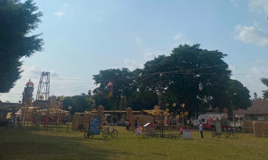 Open-air park festival with hay sculptures, hanging decorations, and people strolling under a large tree on a sunny day.