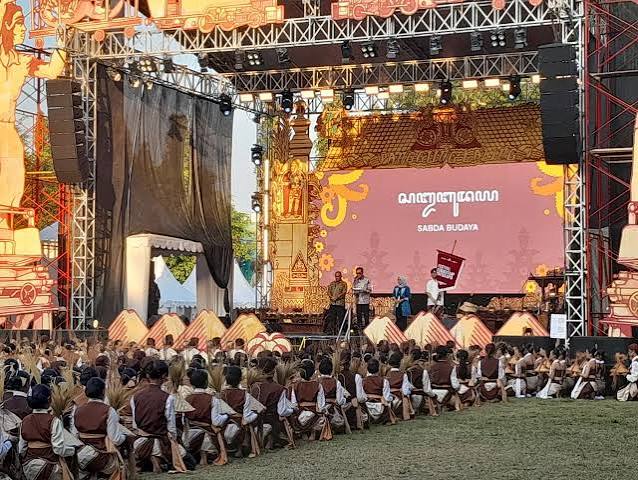 Outdoor cultural stage with performers; crowd seated on grass in traditional brown-and-white attire; large screen displays Malayalam text.