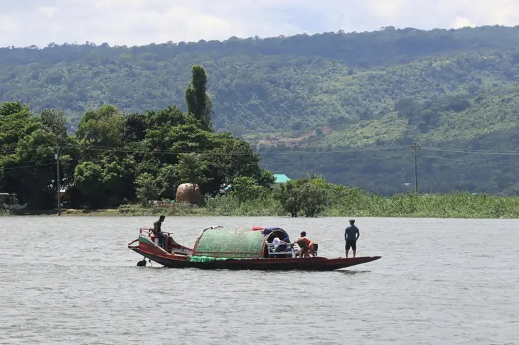 People stand on a small boat with a green woven shelter sailing on a wide river, with forested hills in the background.